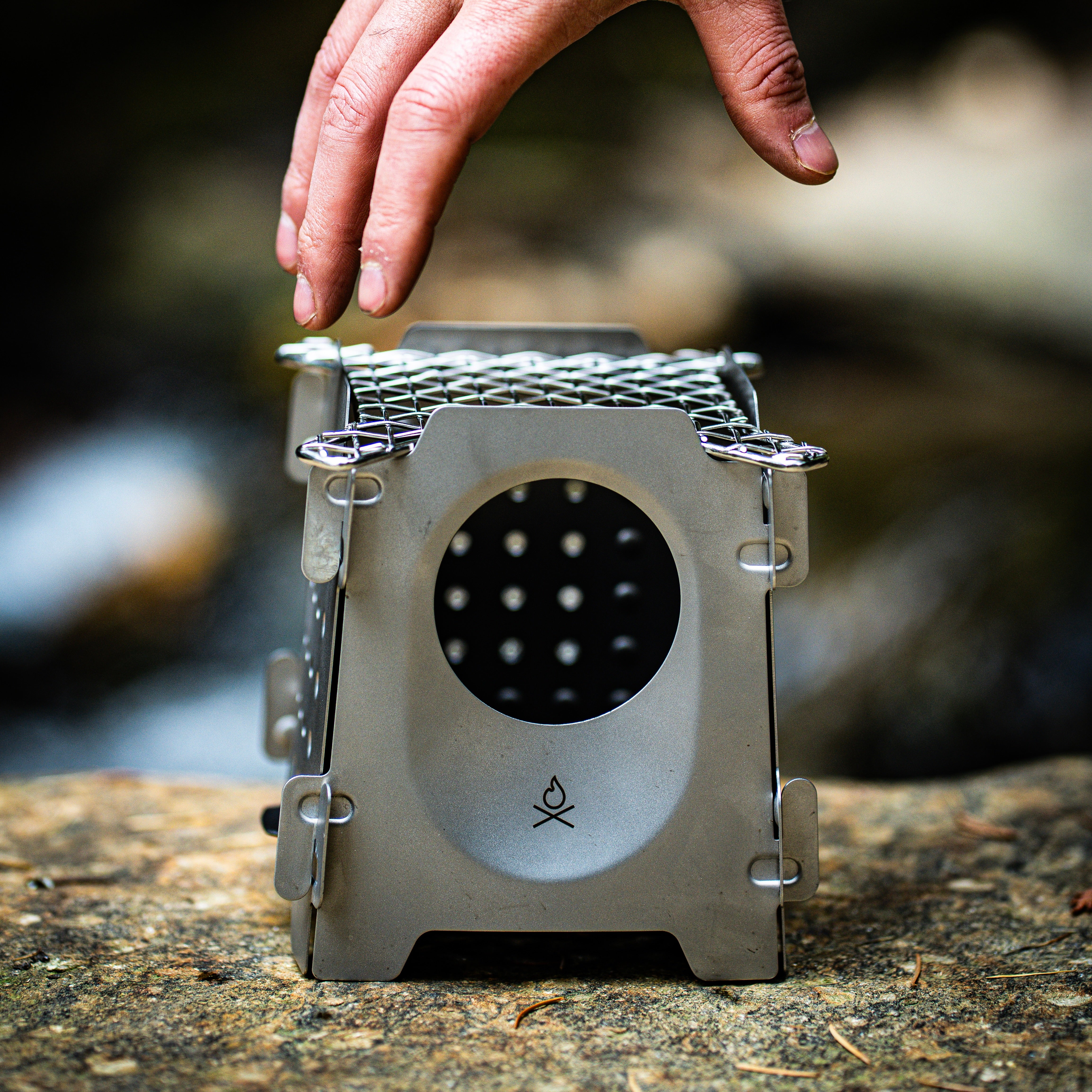A hand is seen placing a small, stainless steel twig stove with a circular opening, on a rocky surface. The Überleben Stöker Stove which uses organic fuel, features a simple fire icon on the front.