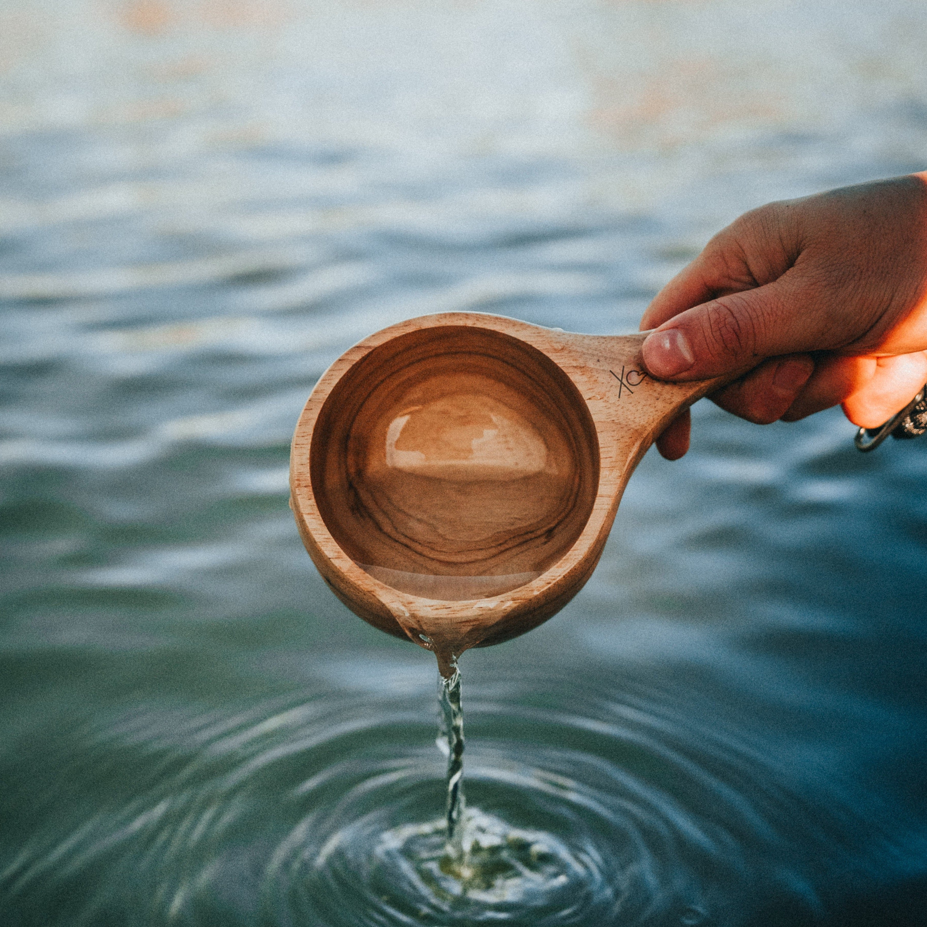 A hand holds the Lore Kuksa by Überleben, scooping water from a lake. This handcrafted kuksa, made from 100% natural hardwood, boasts a smooth, polished finish.