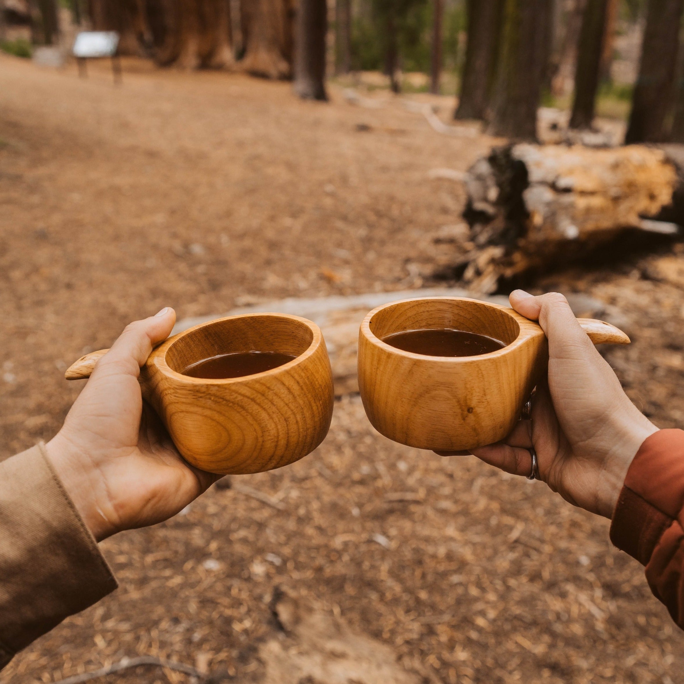 Two hands holding hand-carved Lore Kuksa cups by Überleben, filled with cofffee, in a forest setting.