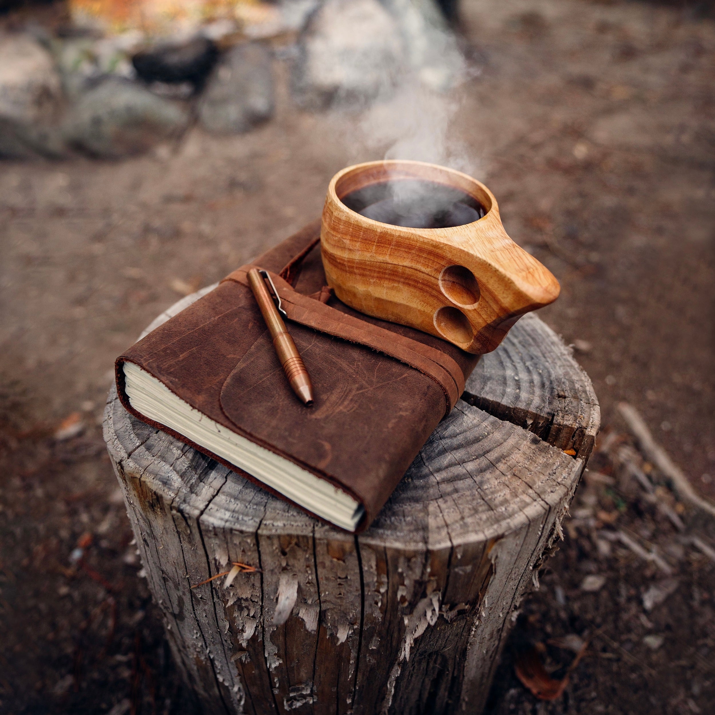 The Original Kuksa camp cup from Überleben, filled with steaming hot coffee, sits atop a closed leather-bound journal. This setup sits on top of a tree stump against a backdrop of blurred rocks and nature.