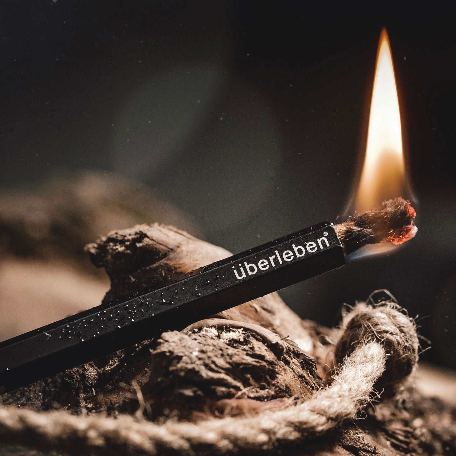 A lit fire starter, the Tindår Wick & Bellow Kit. Branded "Überleben," its ultralight weatherproof paraffin-wax infused hemp wick burns steadily at one end, surrounded by dried leaves that suggest an outdoor setting.