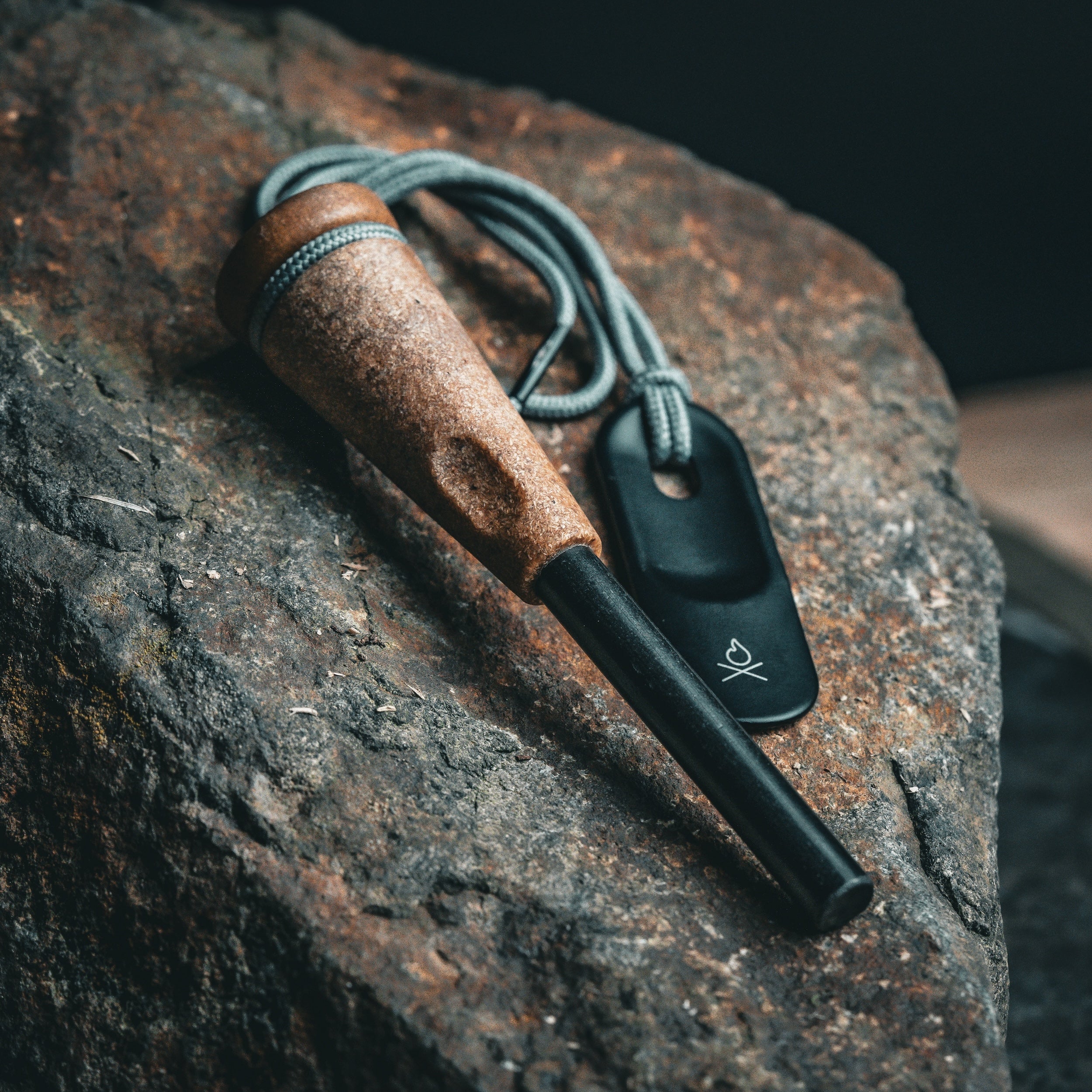 An Überleben fire starter, the Zünden 2.0, features a modern design, and includes a wood composite handle and a black ferro rod that perfectly complements the rugged dark brown rock it is resting on. It also comes with a gray paracord loop attached to the fire starter, which is connected to a black metal striker showcasing a fire logo.