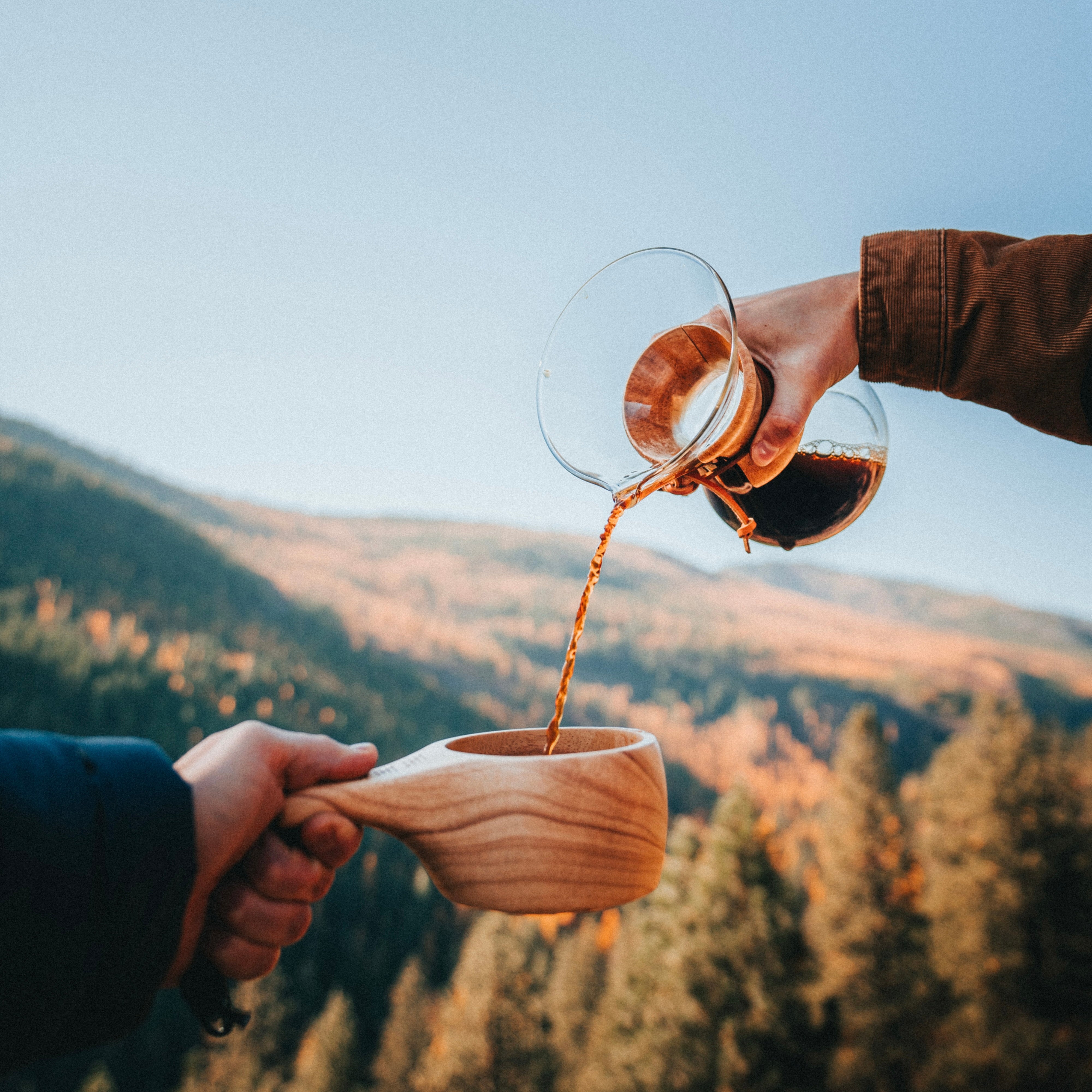 A hand pours coffee from a glass coffee maker into an Überleben Lore Kuksa, with the background showcasing a scenic view of a dense forest under a clear sky.