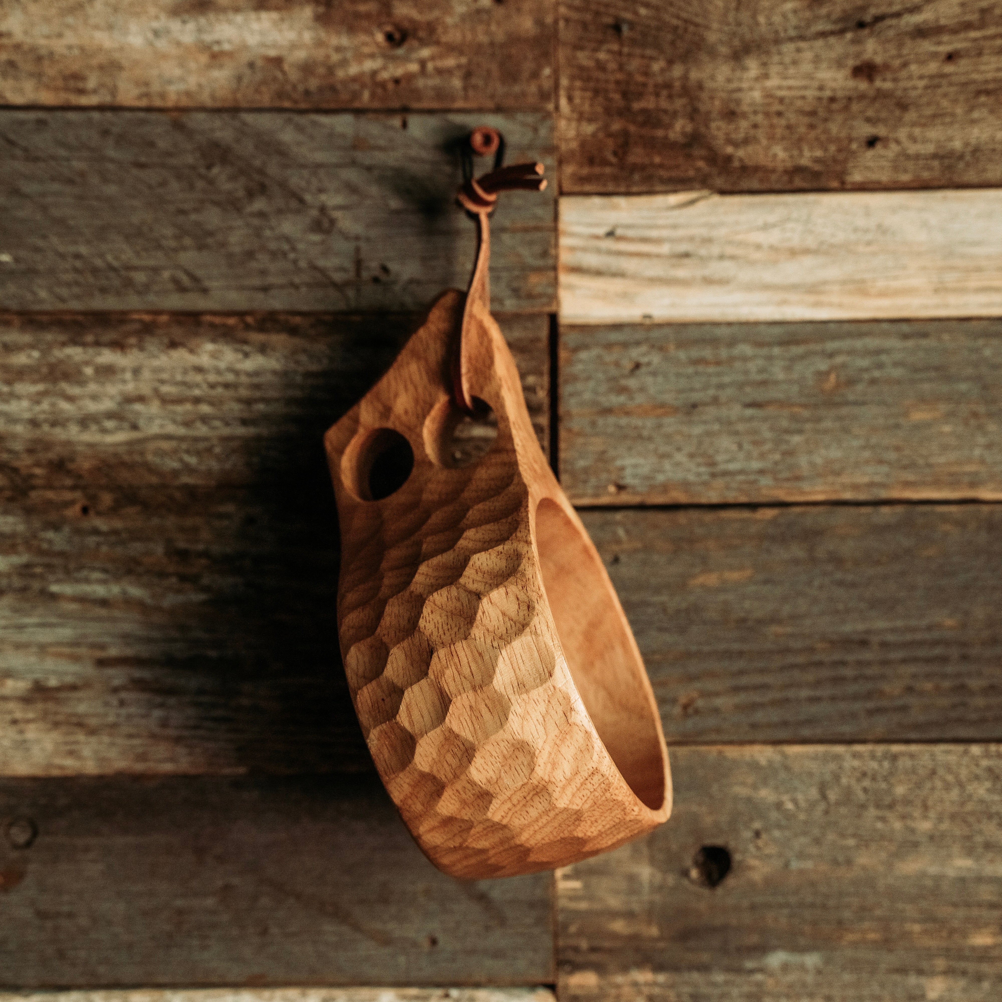 The Überleben Original Kuksa, a handcrafted camp cup made from natural hardwood and featuring a carved design with a handle that has two holes, hangs from a leather rope on a rustic wooden wall. Its Nordic design adds to the charm of the setting.