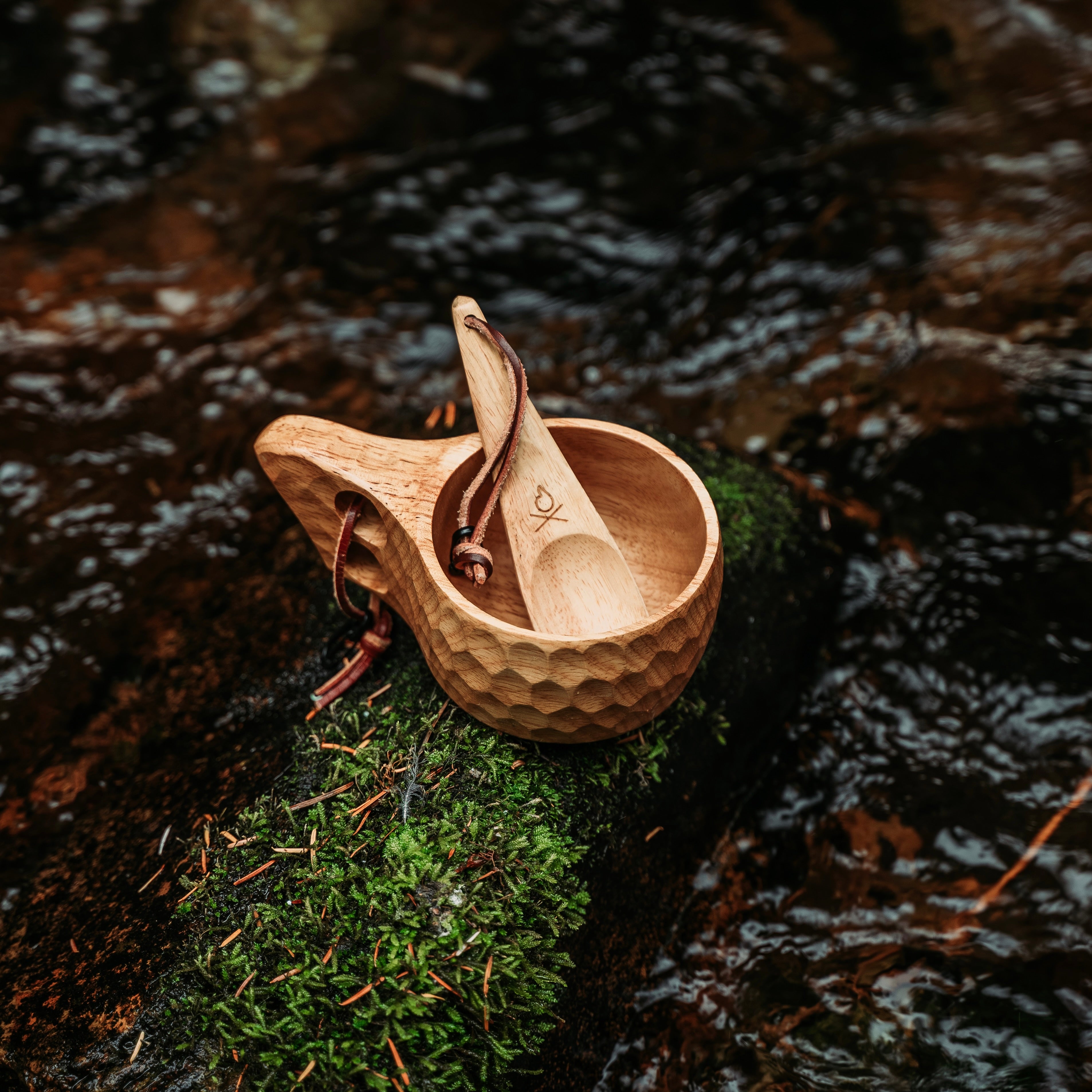 An Original Kuksa camp cup from Überleben and a spoon rest on a moss-covered rock next to a flowing stream. The natural hardwood cup flaunts a honeycomb pattern and a looped leather strap attached to the handle, while the spoon is nestled inside, showcasing Nordic design.