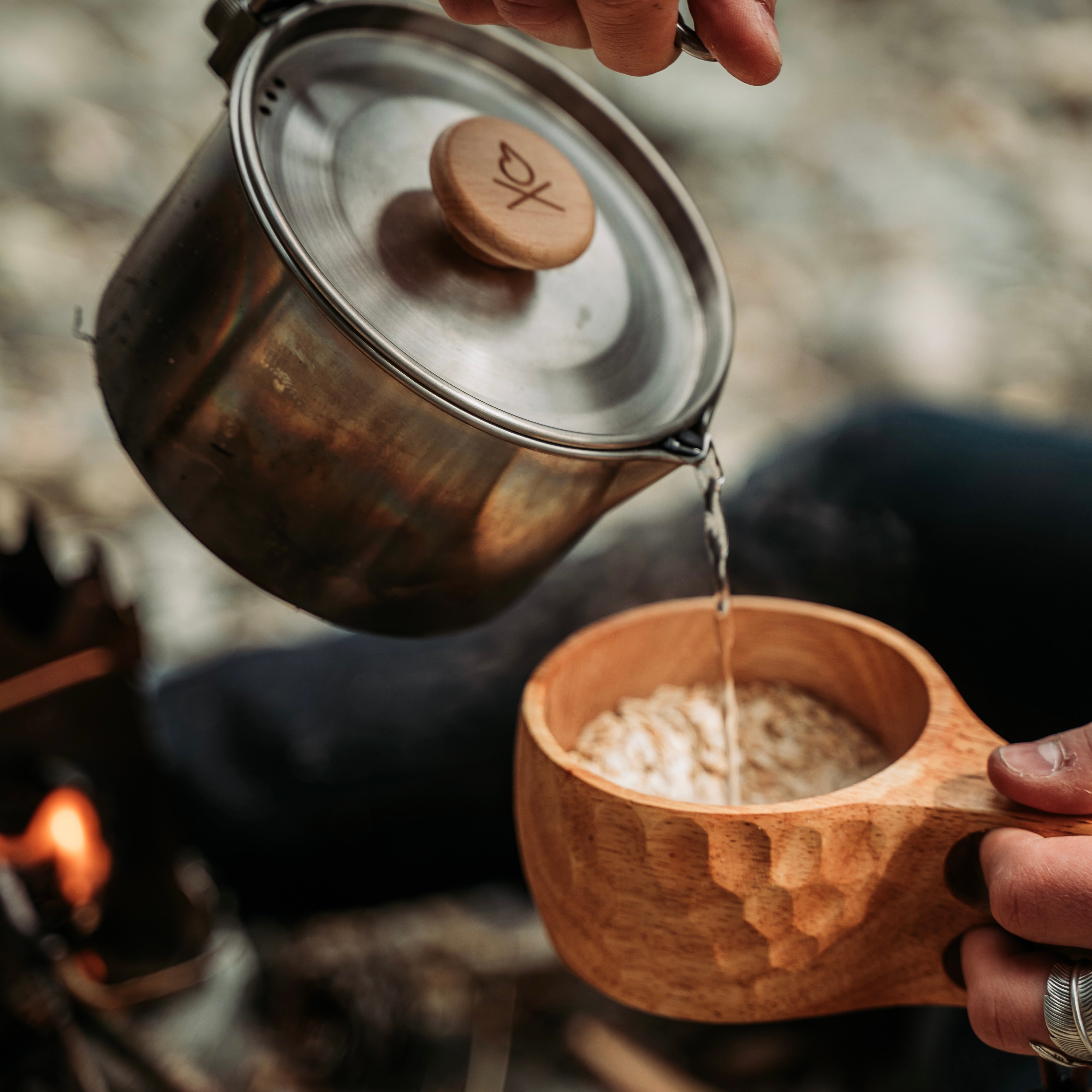 A person is pouring hot water from a metal pot into Überleben's handcrafted Original Kuksa camp cup, which is filled with oats. The natural hardwood of the Original Kuksa highlights its Nordic design, enhancing the peaceful outdoor scene.